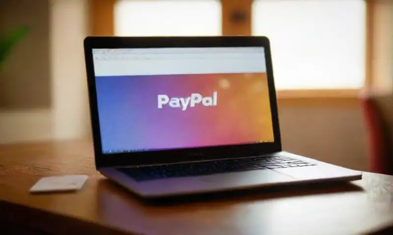 Un portátil elegante brilla en una mesa de madera, mostrando el logo de PayPal, rodeado de cartas y dados en un ambiente acogedor y bien iluminado